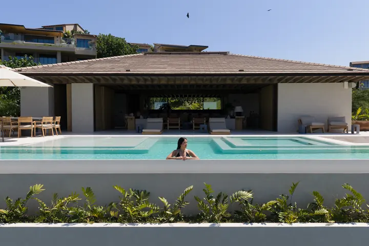 The pool before the open-sided living pavilion, with loungers, a table under an umbrella and a swimmer at the edge