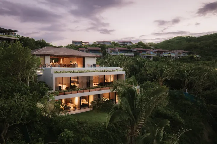The three floors of Villa Taiyari lit at dusk above the lawn, neighbouring homes on the wooded hillside behind