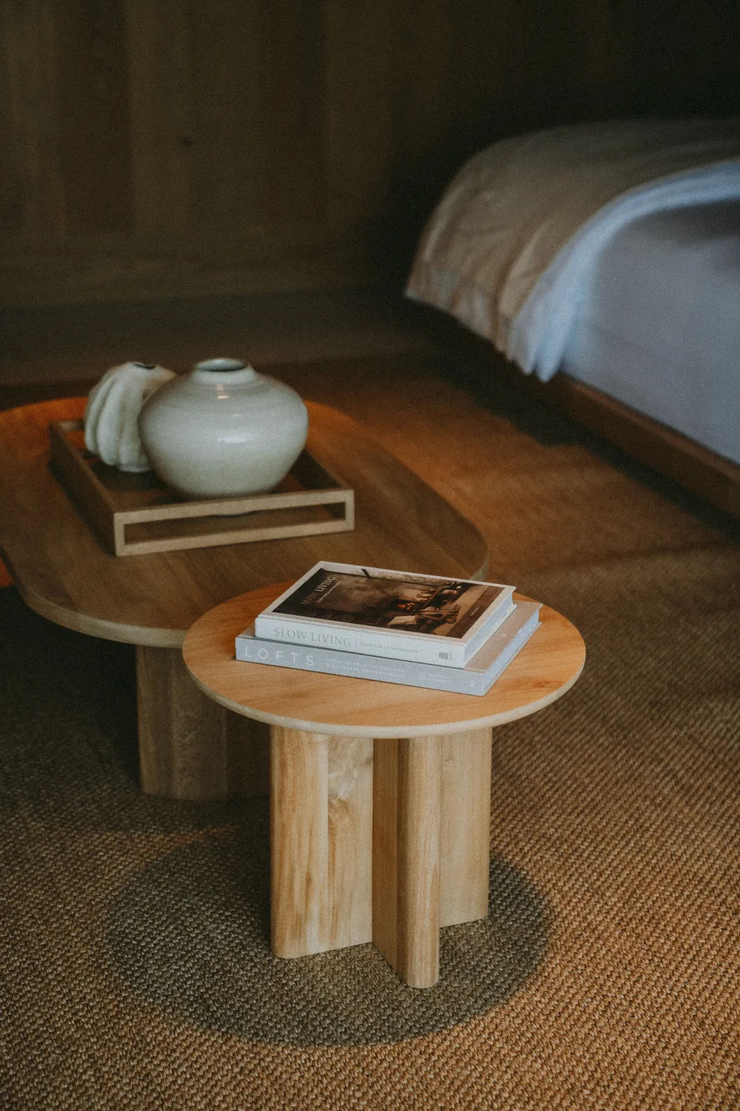A round wooden side table with books and a ceramic vase beside a made bed on a woven rug