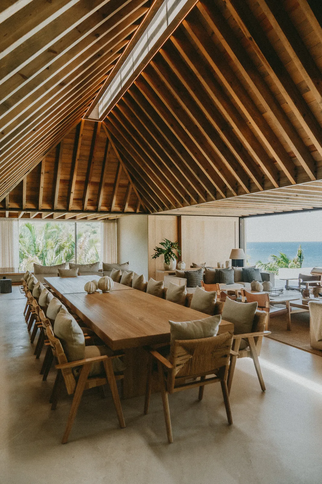 The great room under its peaked timber roof: a long wooden dining table, sofas beyond and the ocean through the glass