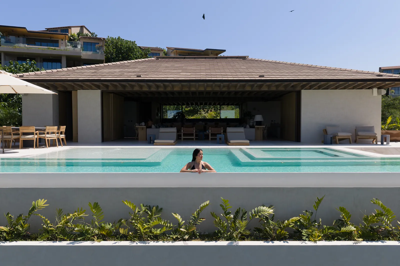 The pool before the open-sided living pavilion, with loungers, a table under an umbrella and a swimmer at the edge