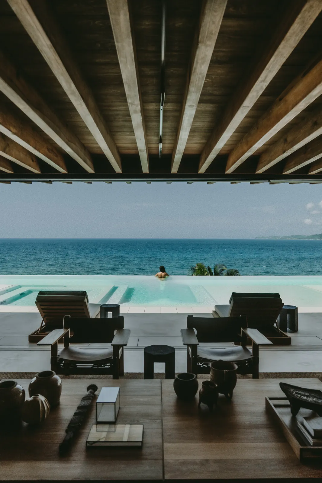 The view from under the timber beams: armchairs, the infinity pool and the open Pacific, a swimmer at the far edge